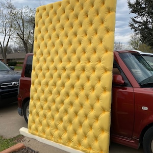 May include: A large, rectangular, tufted headboard in a vibrant yellow color. The headboard is supported by a white base and is leaning against a red car. The tufting creates a diamond pattern across the surface.