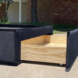 May include: A black upholstered storage bed frame with a wooden drawer that is pulled out. The drawer is on wheels and is made of plywood.