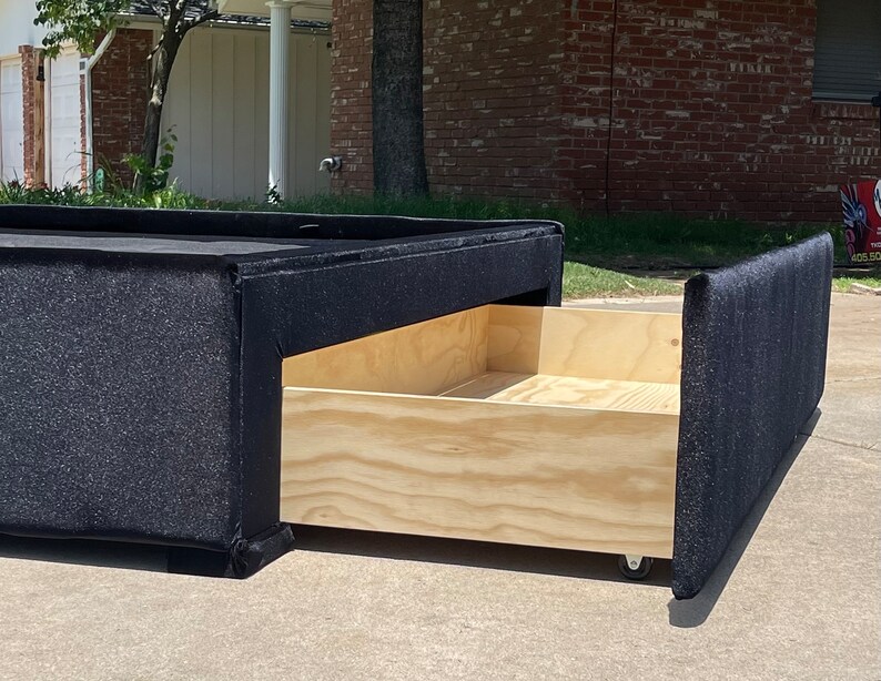 May include: A black upholstered storage bed frame with a wooden drawer that is pulled out. The drawer is on wheels and is made of plywood.