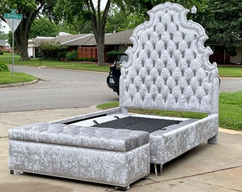 May include: A silver velvet upholstered bed frame with a tufted headboard and a matching storage ottoman. The headboard features a decorative, ornate design.