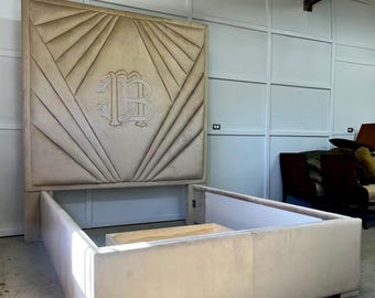 Puede incluir: Un marco de cama tapizado beige con cabecero alto. El cabecero presenta un diseño geométrico con un monograma central. El marco de la cama es rectangular y bajo.
