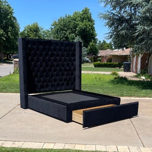 May include: A black upholstered bed frame with a tufted headboard and a pull-out drawer. The bed frame is on a concrete driveway with green grass in the background.