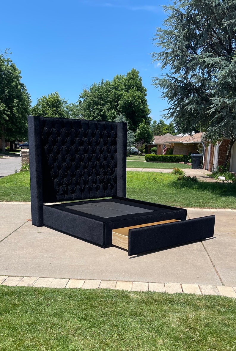 May include: A black upholstered bed frame with a tufted headboard and a pull-out drawer. The bed frame is on a concrete driveway with green grass in the background.
