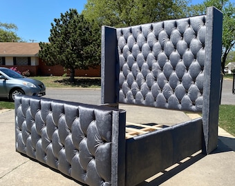Puede incluir: Un armazón de cama tapizado gris con cabecero y piecero altos y con mechones. El armazón de la cama está hecho de un material suave y aterciopelado y tiene un diseño moderno. El armazón de la cama está sobre una superficie de hormigón.