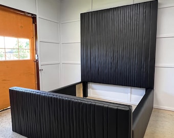 Puede incluir: Un moderno marco de cama negro con una cabecera alta con líneas verticales. El marco de la cama presenta un diseño de perfil bajo con líneas verticales a juego en el pie de cama y los laterales. La cama está contra una pared blanca.