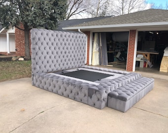 Puede incluir: Un marco de cama tapizado gris con cabecera y compartimentos de almacenamiento. El marco de la cama está hecho de madera y tiene un diseño capitoné. El marco de la cama está en un garaje.