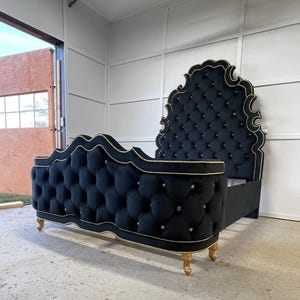 Puede incluir: Un marco de cama de terciopelo negro con cabecero y piecero acolchados. El marco tiene ribetes dorados y patas doradas decorativas. El cabecero tiene una forma ornamental única.