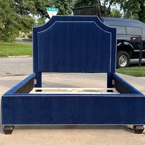 May include: A navy blue upholstered bed frame with a tall, arched headboard. The bed frame has a white trim and sits on dark wooden feet. The bed frame is empty, ready for a mattress.