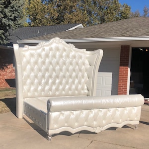 May include: A white upholstered bed frame with a tall, tufted headboard and a rolled footboard. The headboard features a decorative design at the top and the footboard has a scalloped edge.