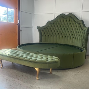 Op de afbeelding: Een luxe olijfgroen, rond bed met een gecapitonneerd hoofdbord en voet. Een bijpassende bank met goudkleurige poten staat voor het bed. Het bed en de bank zijn bekleed met een zachte, fluweelachtige stof.