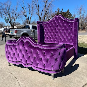 Puede incluir: Un cabecero de cama grande de terciopelo morado con un cabecero y un piecero capitoné. El cabecero de cama tiene un diseño decorativo y ornamentado y patas plateadas.