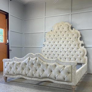 May include: A luxurious, upholstered bed frame with a tufted headboard and footboard. The bed features a light beige fabric with button detailing and a curved design. The frame rests on wooden legs, creating an elegant and classic aesthetic.