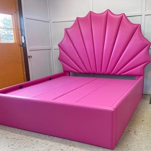 Puede incluir: Un marco de cama tapizado de color rosa con una cabecera única en forma de concha. La cabecera está hecha de múltiples capas de tela, creando un diseño en forma de abanico.