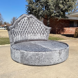 Puede incluir: Un marco de cama redondo de color gris con capitoné y una cabecera grande y adornada. La cabecera está cubierta con una tela plateada con un diseño capitoné.