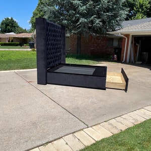 May include: Black upholstered bed frame with a tufted headboard and a pull-out storage drawer. The bed frame is on a concrete driveway.