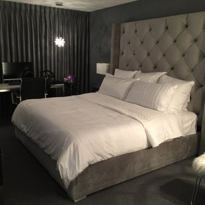 Puede incluir: Un dormitorio lujoso con una cama grande, un cabecero acolchado y ropa de cama blanca. La habitación tiene cortinas grises oscuras, un escritorio con una computadora y una mesita de noche con una lámpara. La estética general es moderna y elegante.