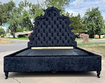 May include: A black velvet bed frame with a tufted headboard. The headboard has an ornate, curved design. The bed frame sits on four carved legs. The bed is outdoors on a concrete surface.