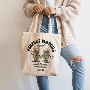K&ouml;nnte beinhalten: Cremefarbene Tragetasche mit den Worten "Perfect Matcha" in einem kreisf&ouml;rmigen Design. Die Tasche zeigt eine Cartoon-Illustration von zwei Matcha-Getr&auml;nken mit Gesichtern, Strohhalmen und Armen. Darunter steht "Fresh Matcha served daily COFFEE."