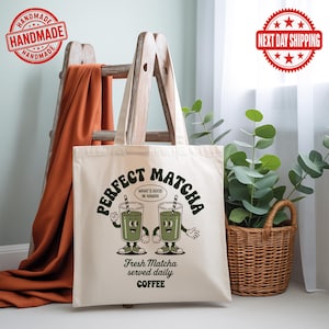 K&ouml;nnte beinhalten: Cremefarbene Tragetasche mit dem Schriftzug "PERFECT MATCHA" in Retro-Schrift und zwei Cartoon-Matcha-Getr&auml;nken. Der Text "WHAT'S GOOD IN HAWAII" ist ebenfalls vorhanden. Auf der Unterseite der Tasche steht "Fresh Matcha served daily COFFEE".