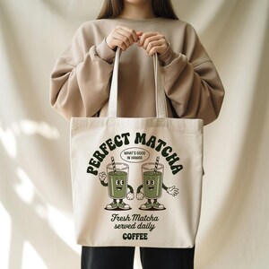 K&ouml;nnte beinhalten: Cremefarbene Tragetasche mit dem gr&uuml;nen Schriftzug "PERFECT MATCHA" und Cartoon-Matcha-Getr&auml;nken. Der Text "WHAT'S GOOD IN HAWAII" befindet sich in einer Sprechblase. Auf der Unterseite der Tasche steht "Fresh Matcha served daily COFFEE".