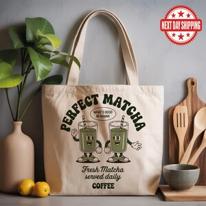 K&ouml;nnte beinhalten: Cremefarbene Tragetasche mit dem gr&uuml;nen Schriftzug "PERFECT MATCHA" und Cartoon-Matcha-Getr&auml;nken. Der Text "What's good in Hawaii" ist ebenfalls vorhanden. Auf der Unterseite der Tasche steht "Fresh Matcha served daily COFFEE".