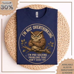 May include: A navy blue t-shirt featuring a circular graphic of a brown owl perched on a branch with its wings crossed. The text reads I'M NOT OVERTHINKING I'M PRE-SOLVING PROBLEMS THAT DON'T EXIST YET in a circular yellow font around the owl.