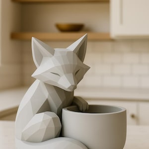 May include: A light gray fox-shaped planter with a geometric design. The fox is seated, with its paws resting on a round, gray planter pot. The planter is on a round base. The fox has closed eyes, and the overall aesthetic is modern.