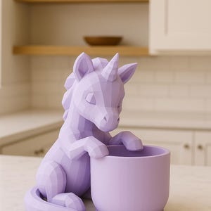 Op de afbeelding: Een lavendelkleurige eenhoorn-bloempot met een geometrisch ontwerp. De eenhoorn zit met zijn voorpoten op de rand van een bijpassende ronde pot. De pot staat op een wit aanrecht in een keuken.