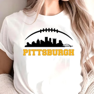 Könnte beinhalten: Weißes T-Shirt mit einer schwarzen Silhouette einer Stadtlandschaft unter einer Footballform. Das Wort "PITTSBURGH" ist in goldenen Buchstaben unter der Skyline aufgedruckt. Das Shirt ist aus weichem Material.