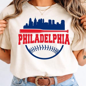 May include: Cream-colored t-shirt featuring a graphic design with the word "PHILADELPHIA" in red, a baseball bat, and a baseball. The design includes a blue skyline silhouette and a baseball stitch detail.