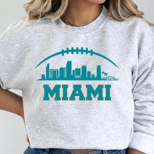 May include: A light grey sweatshirt with a teal graphic of the Miami skyline beneath a football shape. The word "MIAMI" is printed in teal block letters below the skyline design. The sweatshirt has a crew neck.
