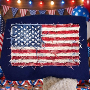 Könnte beinhalten: Ein marineblaues Kissen mit einem abgenutzten Design der amerikanischen Flagge. Die Flagge hat ein denimblaues Quadrat mit weißen Sternen und rot-weiß gestreiften Abschnitten, was ihr einen Vintage- und patriotischen Look verleiht. Dekoriert mit Dekorationen zum 4. Juli.