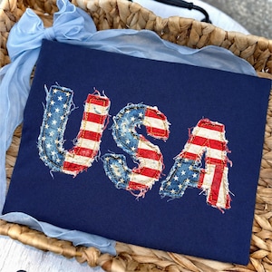 May include: A navy blue shirt with the word "USA" appliqued in a distressed American flag design. The letters are red, white, and blue, with a frayed edge. The shirt is displayed in a woven basket with a light blue ribbon.