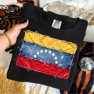 Puede incluir: Sudadera negra con un diseño desgastado de la bandera venezolana. La bandera está hecha de tela amarilla, azul y roja con estrellas blancas. La sudadera tiene cuello redondo y mangas largas. La etiqueta dice "Comfort Colors".