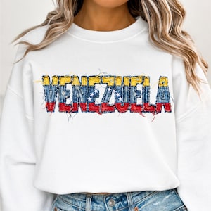 Puede incluir: Sudadera blanca con la palabra "VENEZUELA" en letras con los colores de la bandera venezolana: amarillo, azul y rojo. El diseño es desgastado. La sudadera es de estilo casual y cómodo.