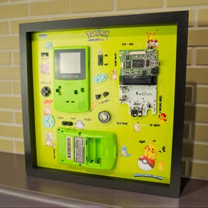 May include: A framed display showcasing a disassembled green Game Boy, with its parts arranged on a lime green background. The display includes the Game Boy's shell, circuit board, and battery compartment, alongside Pokémon character stickers. The frame is black.