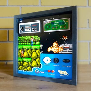 May include: Framed artwork featuring a retro video game scene with pixelated graphics. The artwork includes a controller, game board, and game elements. The color palette includes green, blue, orange, and black.