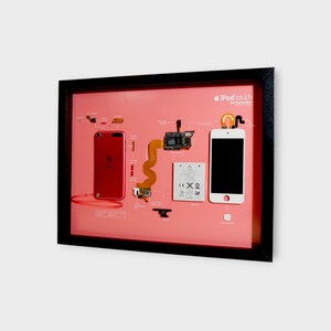 May include: Framed artwork featuring an exploded view of an iPod Touch. The components are arranged on a pink background within a black frame. A red iPod and a white iPod are displayed, along with various internal parts.