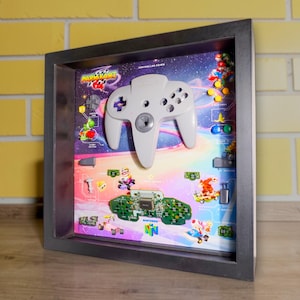 May include: A framed shadow box featuring a white Nintendo 64 controller. The background showcases a colorful scene from the Mario Kart 64 game, with various characters and elements. The frame is dark gray.