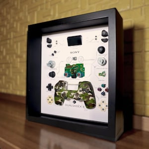 May include: A framed display featuring a deconstructed PlayStation DualShock 4 controller. The components are arranged on a white background, with the controller's camouflage-patterned shell prominently displayed. The frame is black, and the Sony logo is visible.