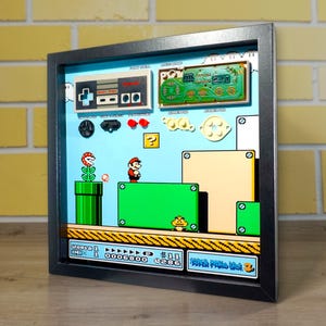 May include: Framed artwork featuring a Super Mario Bros. scene. The artwork includes a controller, circuit board, and a level from the classic video game. The scene shows Mario, green pipes, and question mark blocks. The frame is black.