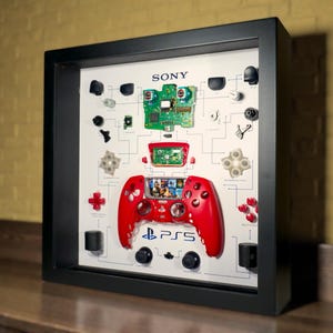 May include: A framed display showing a red PS5 controller and its dismantled parts. The controller is central, with its buttons and internal components arranged around it. The Sony logo and PS5 logo are visible.