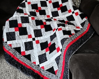 May include: A black, white and red quilt with a geometric pattern. The quilt is folded and draped over a sofa.