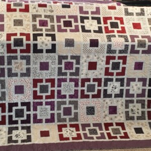 Könnte beinhalten: Eine Steppdecke mit einem geometrischen Muster in Grautönen, Lila und Rot. Die Steppdecke besteht aus Quadraten mit einem Rahmen um jedes.