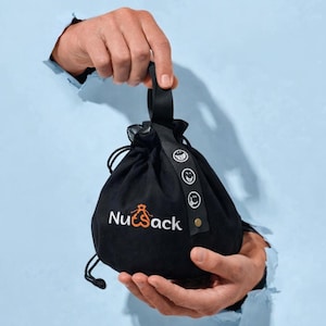 May include: Black drawstring bag with the text "NutSack" in white and an orange graphic. The bag has a black handle and a strap with three smiling face graphics. The bag is held against a light blue background.