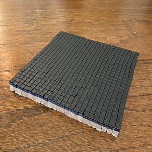 May include: A square, dark gray foam pad composed of many small, individual square blocks. The edges of the pad reveal a white, blue, and gray layered construction. The pad is resting on a wooden surface.