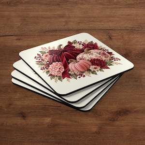 May include: A stack of four square coasters with rounded corners, featuring a cream-coloured background and a central illustration of pumpkins, deep red velvet-textured bows, and clusters of autumn flowers in shades of burgundy and pink.