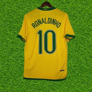 Puede incluir: Una camiseta de fútbol amarilla con ribetes verdes, con el nombre "RONALDINHO" y el número "10" en verde. La camiseta está colgada en una percha de madera sobre un fondo verde.