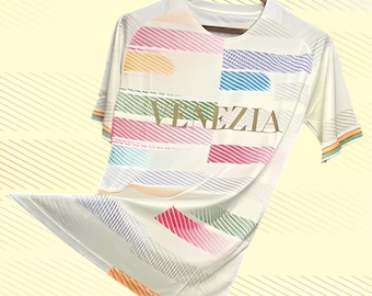May include: Cream-colored t-shirt with a colorful geometric design and the word "VENEZIA" in gold. The shirt features horizontal stripes in various colors, including pink, orange, and blue. The sleeves have a striped trim.