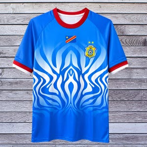 Könnte beinhalten: Ein blaues Fußballtrikot mit einem abstrakten weißen und blauen Design. Das Trikot hat einen roten Kragen und Ärmelbesatz. Eine kleine Flagge und ein Team-Emblem befinden sich auf der Vorderseite.